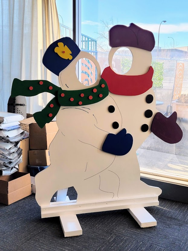 a large wood board cut and painted in the shape of 2 snowmen wearing scarves and hats with the faces cut out.