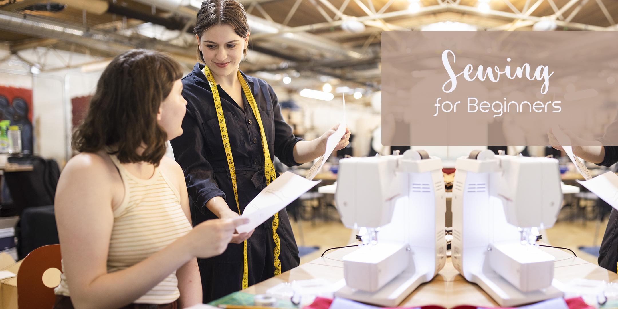 one woman is sitting and one woman is standinh infrotn of a sewing machine. the standing woman has a measuring tape around her neck