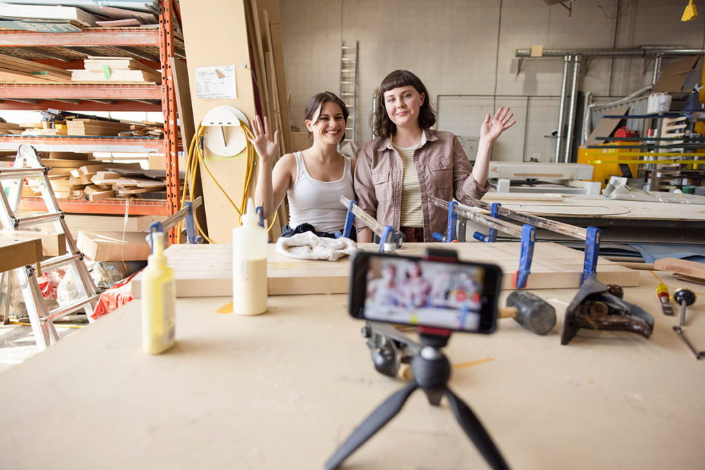 2 people smile an wave in a workshop space in the background while a camera is setup in the foreground filming them. We can see their image in the cameras viewfinder screen