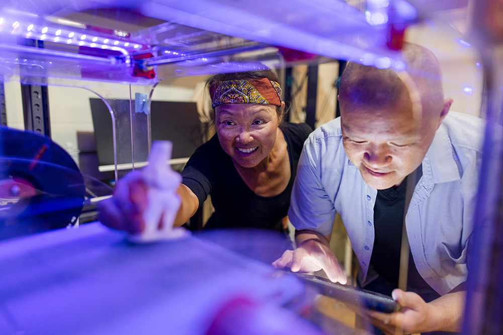 2 people look inside a 3d printer as it prints an object. the printer is lit up with colorful light at Fuse 33 makerspace in calgary.