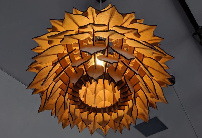 A laser cut wood light with a flower like pattern made of small wood pieces of wood radiating outward from the centre.
