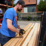 a man in a blue Superman shirt and a baseball hall measures wood at a mitre saw. He is standing outside