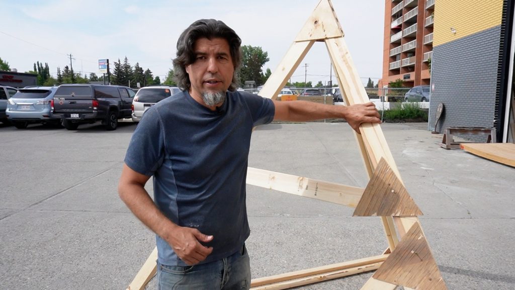 Ken Hack standing beside a large wooden triangle frame-one of many that makes up the shape of the freezer burn 2023 effigy